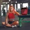 A woman sitting on a bench in a gym using an advanced red light panel called REDi Rejuv.